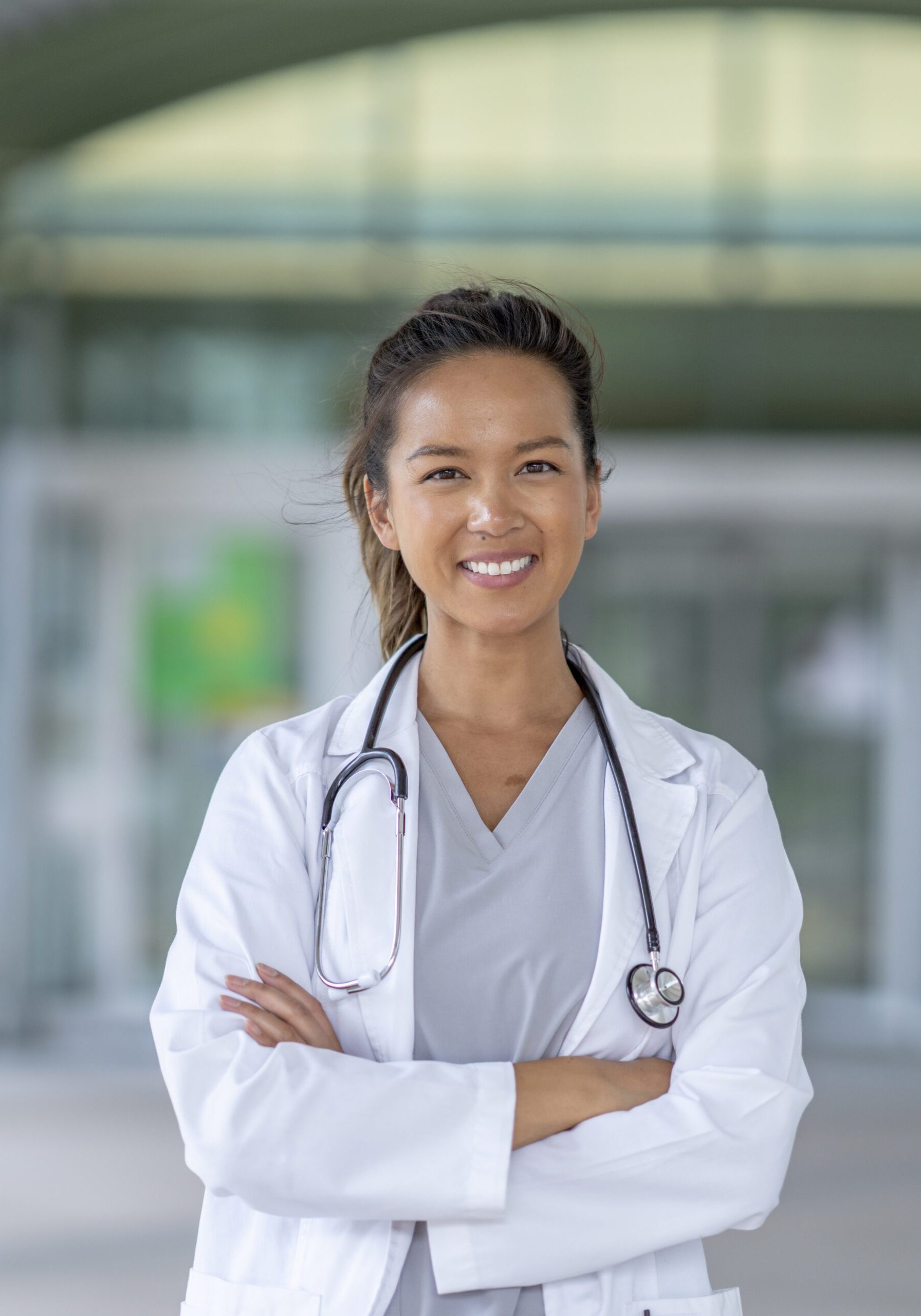 A female doctor of Asian decent, stands outside a hospital as she poses for a portrait.  She is wearing a white lab coat and has a stethoscope around her neck with her arms crossed in front of her as she smiles.